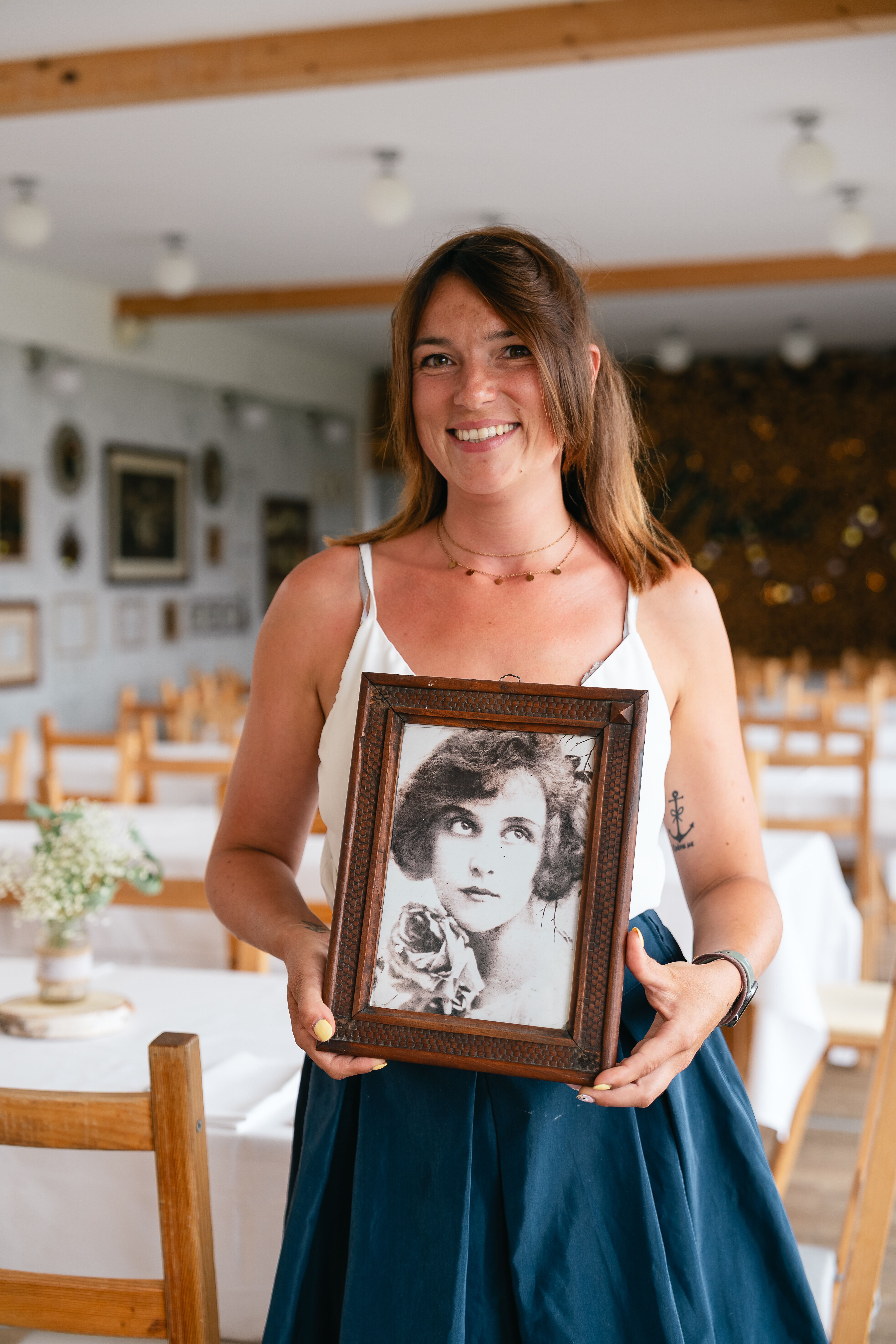 Sarah Trybek steht im lichtdurchfluteten Gastraum des Gasthauses Mirli und hält ein gerahmtes Bild der früheren Wirtin Mirli, zu deren Ehren das Haus benannt wurde. Die Umgebung strahlt eine freundliche und einladende Atmosphäre aus.