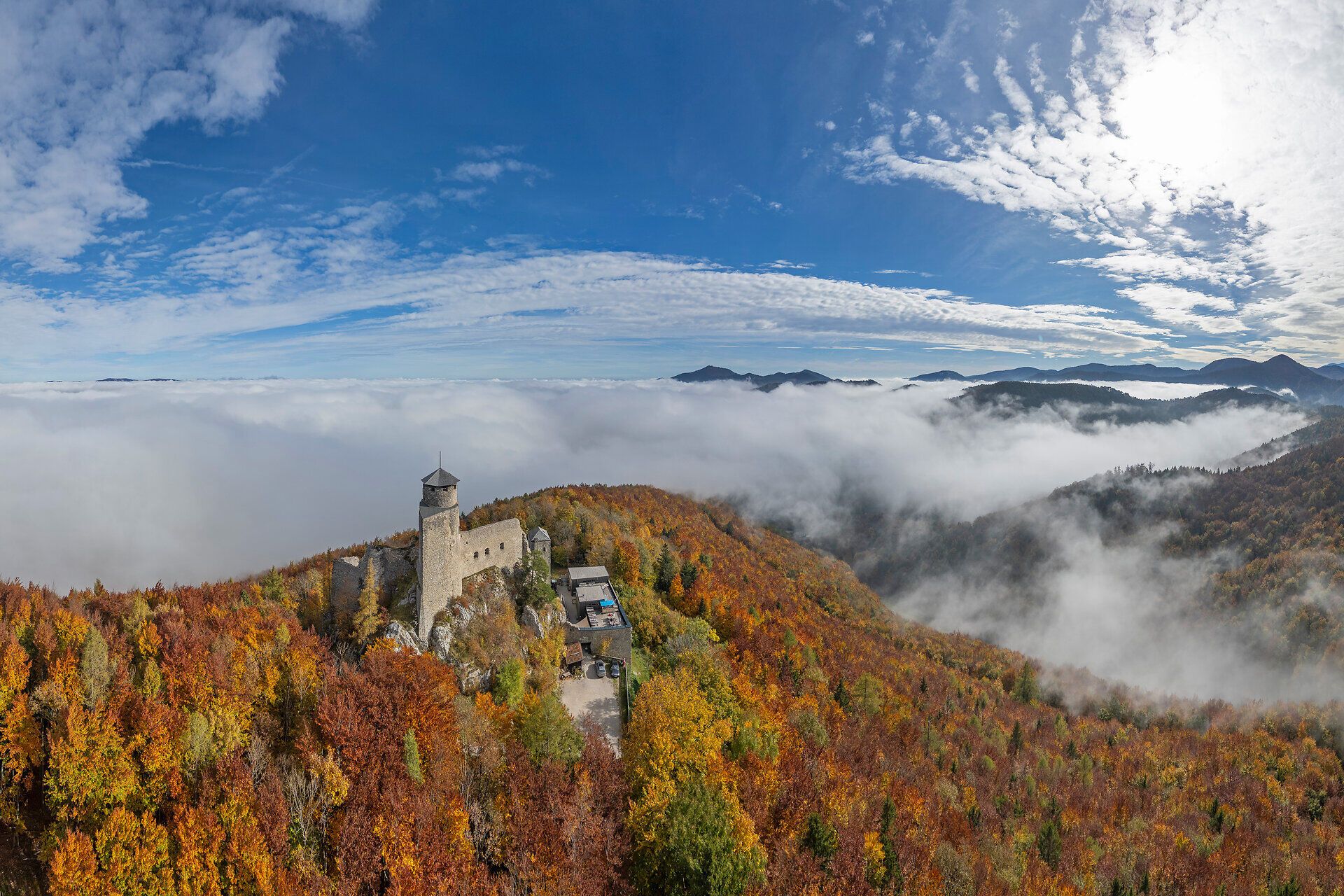 Luftaufnahme der Araburg im Herbst: Die Burg ist von orange- und gelb gefärbtem Wald umgeben, eine dichte Nebeldecke liegt über dem Tal, während oberhalb der Burg strahlender Sonnenschein den Himmel erhellt.