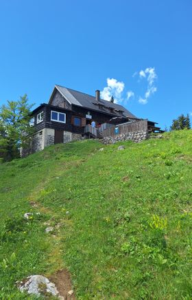 Waldfreundehütte am Obersberg mit Holzverkleidung an einem Hang, davor eine Wiese und blauer Himmel