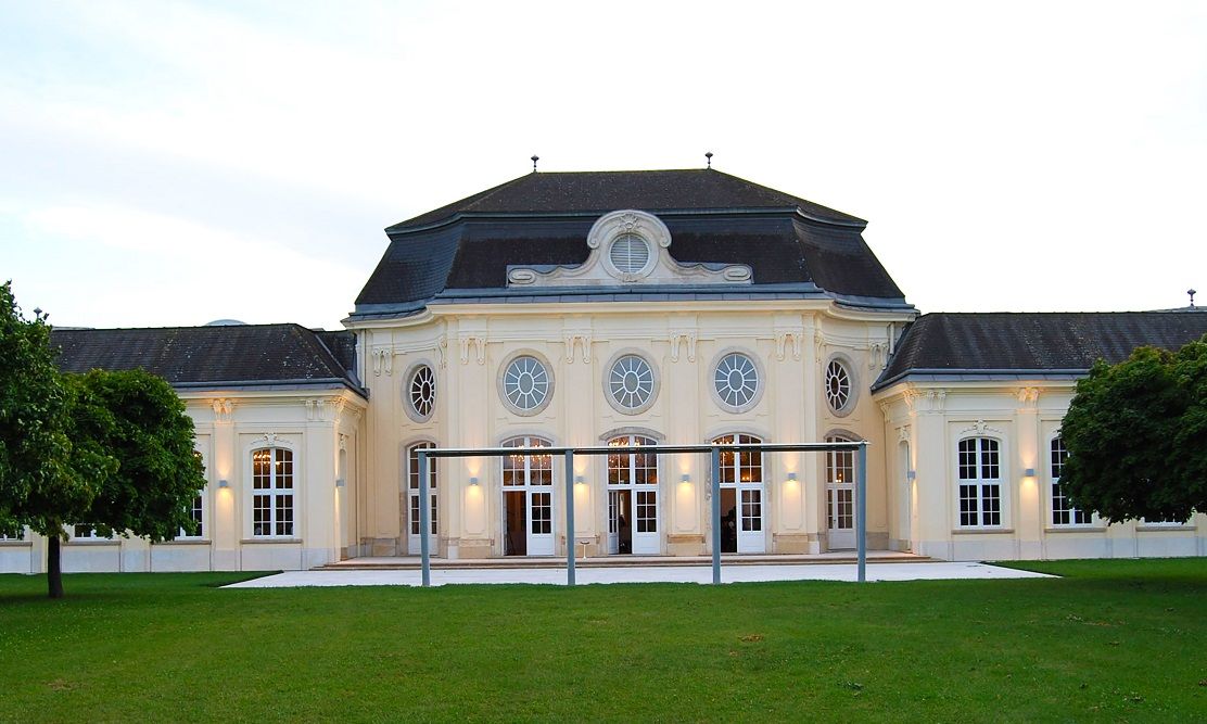 Historisches Gebäude mit symmetrischer Fassade und großen Fenstern, umgeben von Grünflächen.