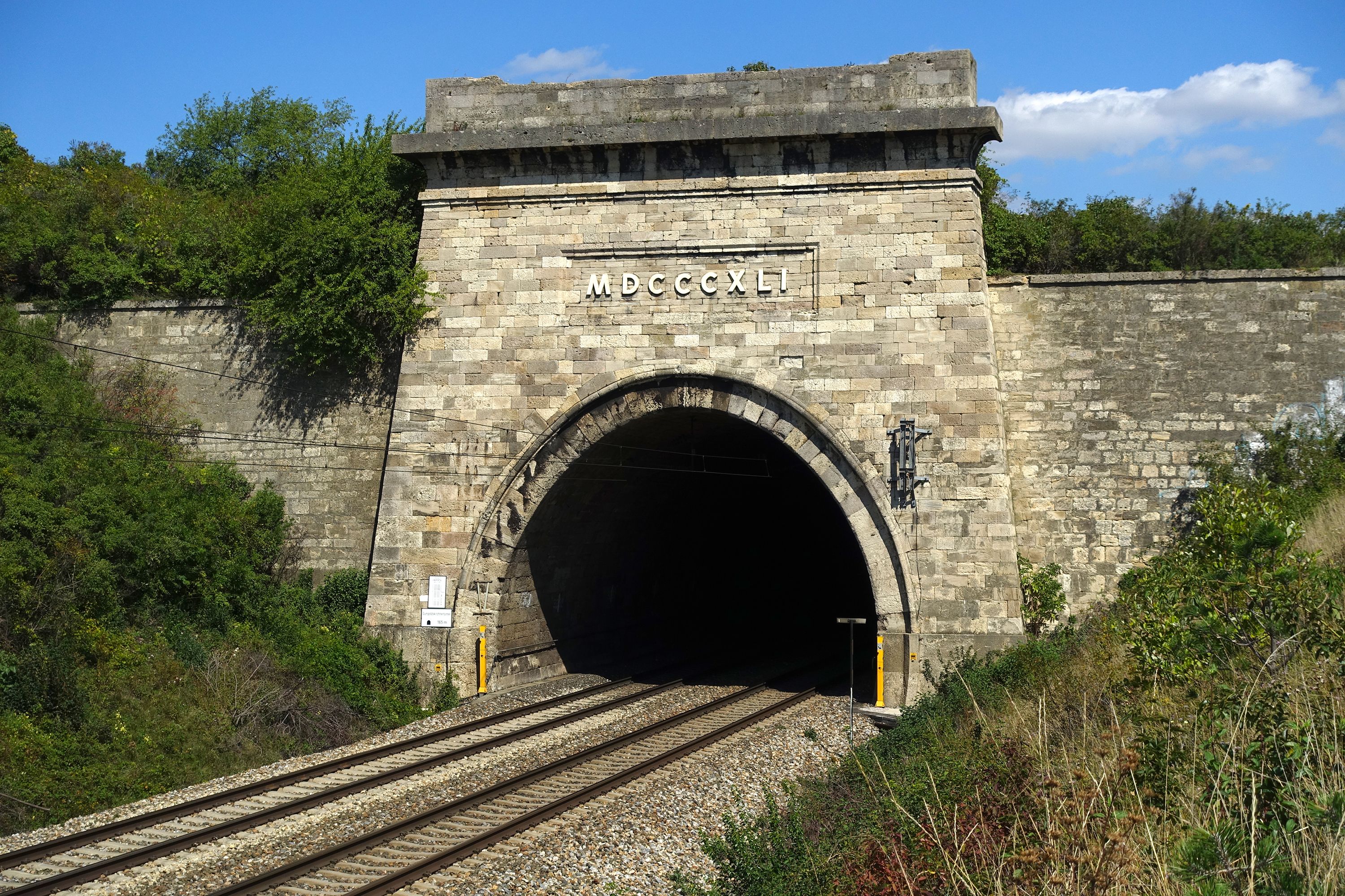 Ein gemauerter Eisenbahntunnel mit rundem Bogeneingang. Vor dem Tunnel verlaufen zwei Bahngleise. Über dem Eingang steht die Jahreszahl 1840 in römischen Ziffern. Rundherum ist grüne Vegetation, der Himmel ist blau mit wenigen Wolken.