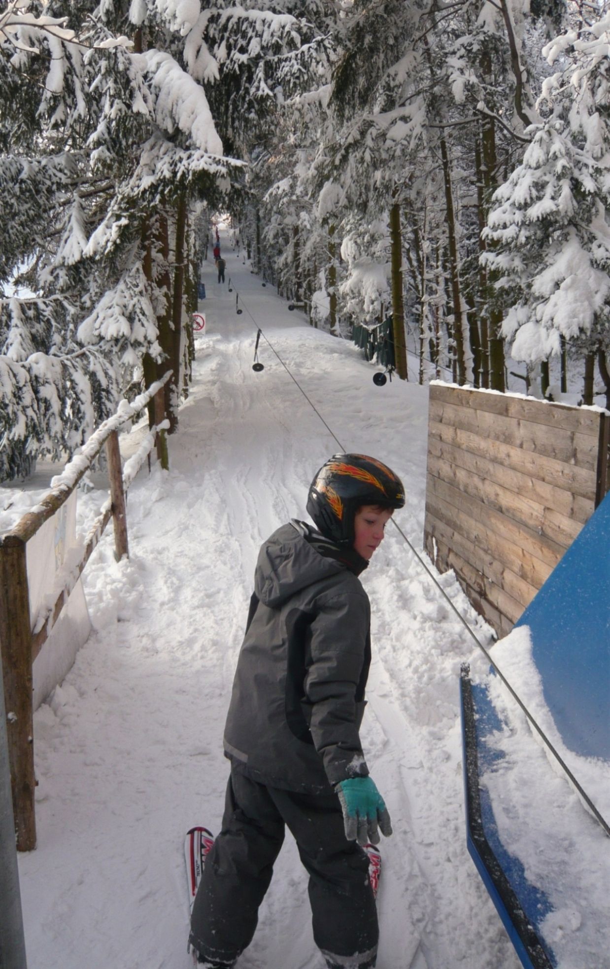 Ein Kind mit Helm steht auf Skiern an einem Schlepplift in einem verschneiten Wald.