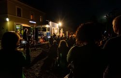 Unter dem strahlenden Licht der Bühne versammeln sich Menschen, um die magische Atmosphäre eines kulturellen Abends zu genießen. Die Klänge der Darbietung vermischen sich mit dem sanften Rascheln der Blätter, während die Nacht langsam hereinbricht und die Umgebung in ein warmes Licht taucht.