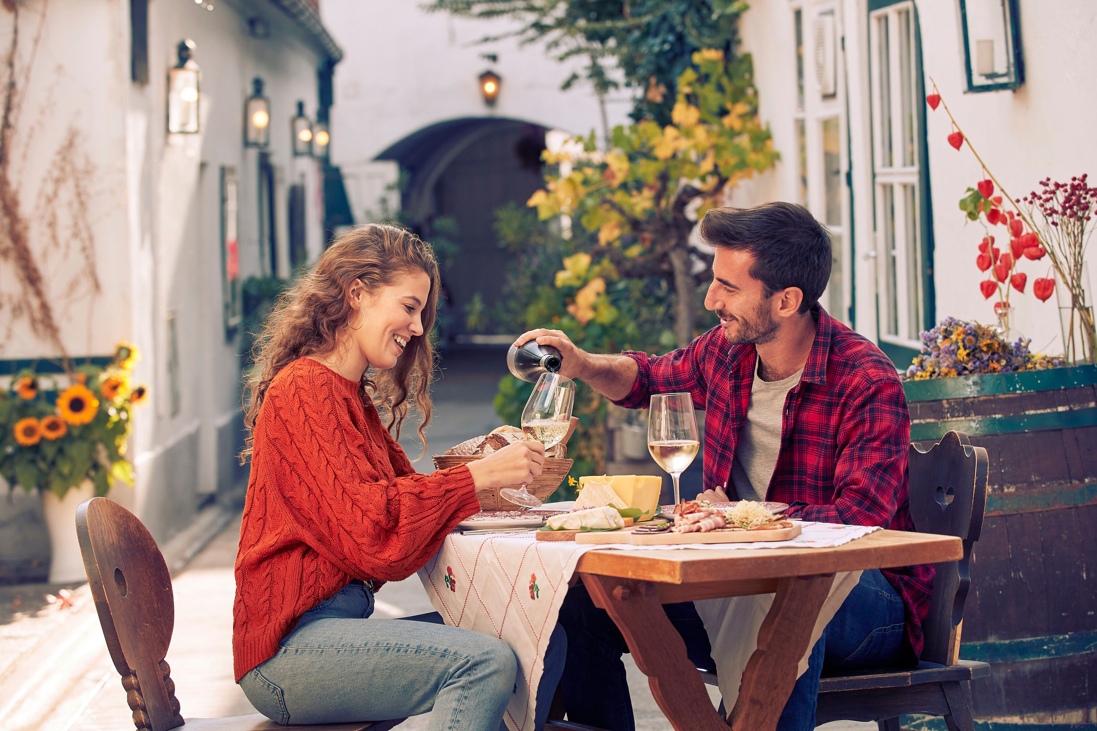Genießen Sie einen romantischen Abend im Freien, umgeben von blühenden Sonnenblumen und der malerischen Kulisse des Triestingtals. Ein Paar teilt sich ein köstliches Essen und ein Glas Wein, während die sanften Farben des Sonnenuntergangs eine warme Atmosphäre schaffen.