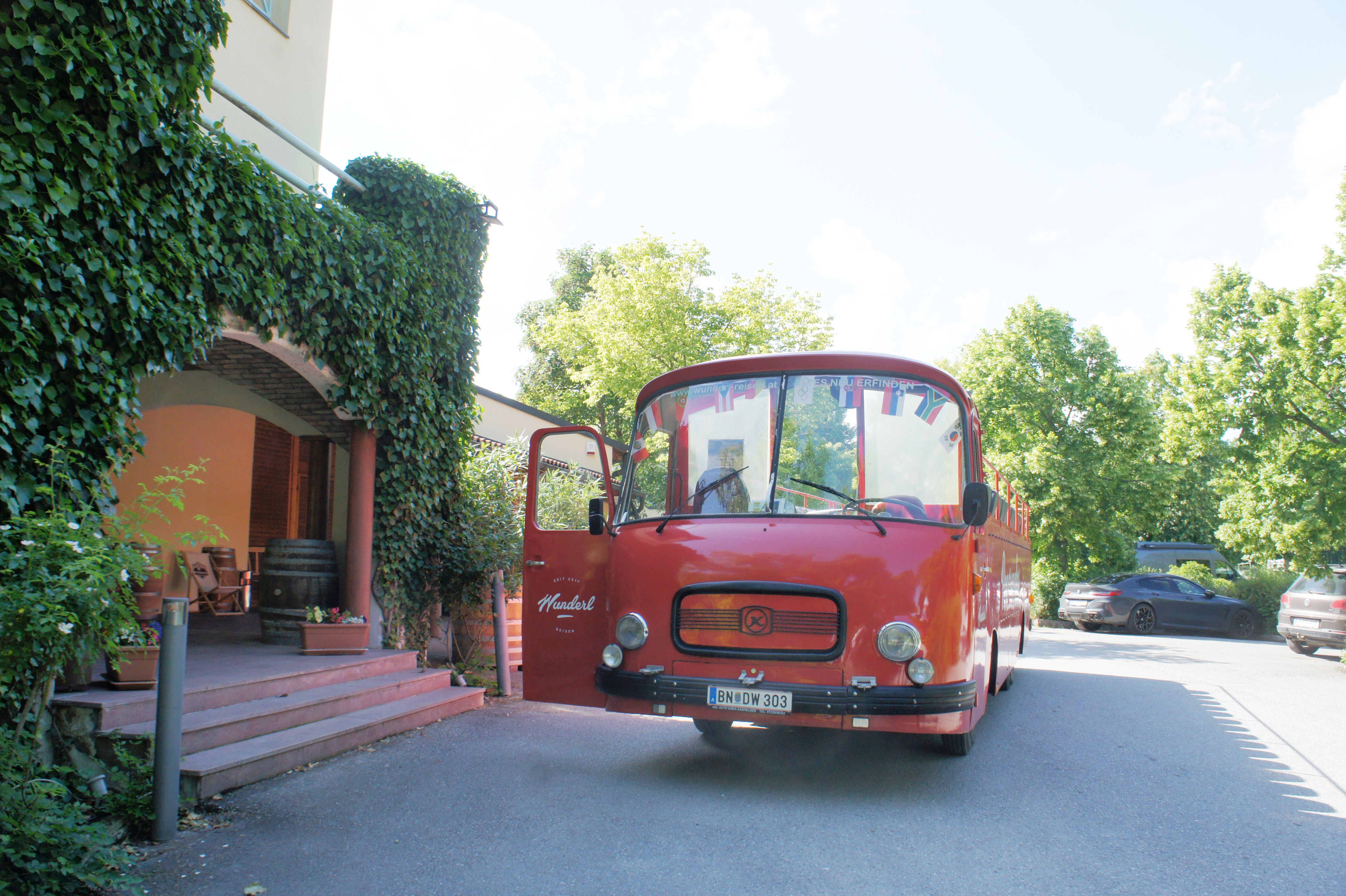 Ein roter Bus mit offenem Oberdeck und Sitzplätzen unter freiem Himmel vor einem Weingut steht geparkt. An der Seite ist die Aufschrift „Panoramabus“ zu sehen. Der Bus wirkt geräumig und lädt zu einer gemütlichen Fahrt mit Aussicht ein.