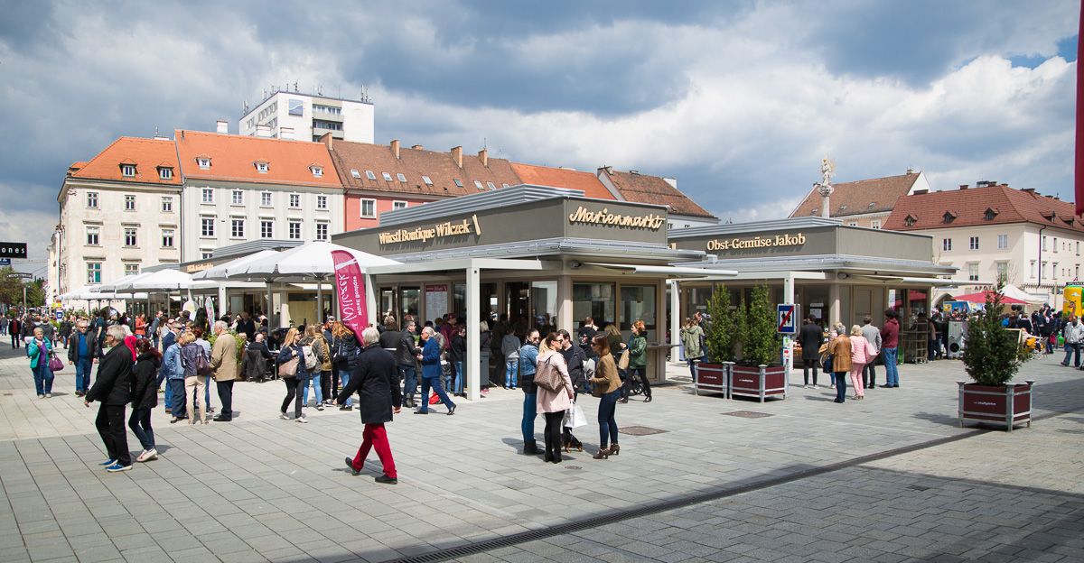 Menschenmenge auf einem belebten Marktplatz mit Ständen und Gebäuden im Hintergrund.