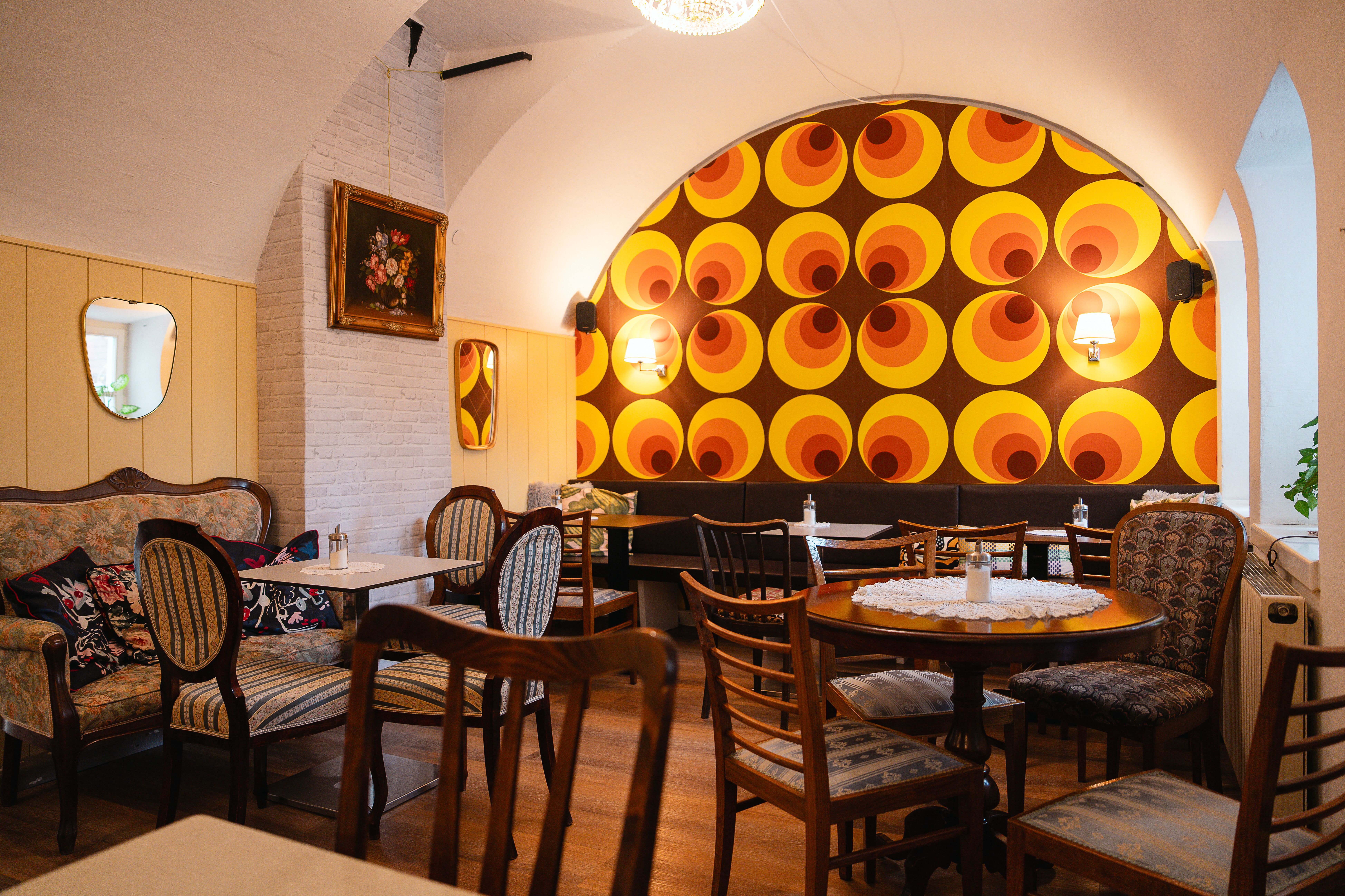 Nostalgisches Kaffeehaus mit Vintage-Charme: Gelb-orange-rote Kreise schaffen ein warmes Willkommen. Dunkle Holztische mit gehäkelten Tischtüchern laden ein, bei Kaffee und Gespräch zu verweilen.