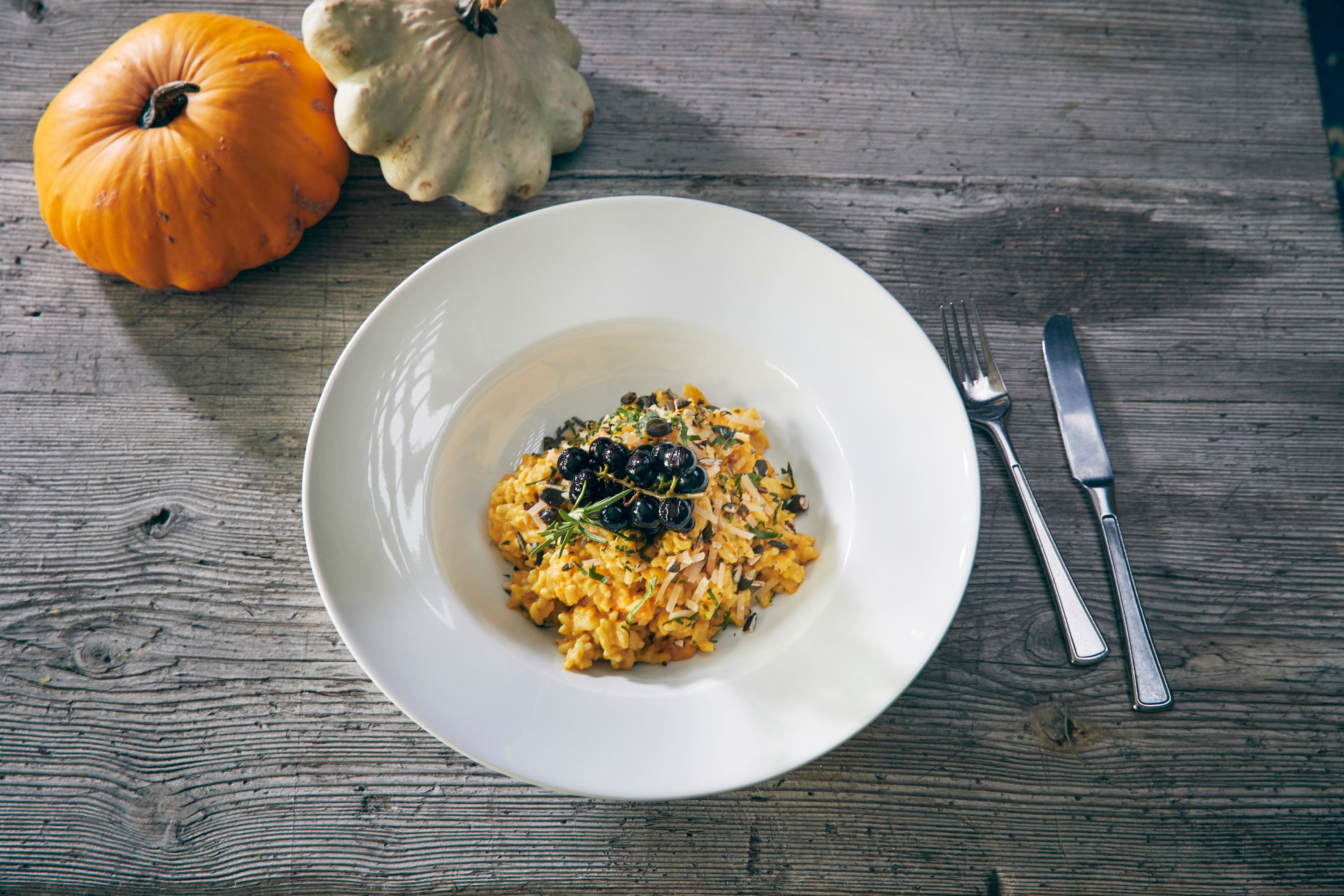 Ein Teller Kürbisrisotto mit Kräutern und Beeren, daneben zwei Kürbisse auf einem Holztisch.