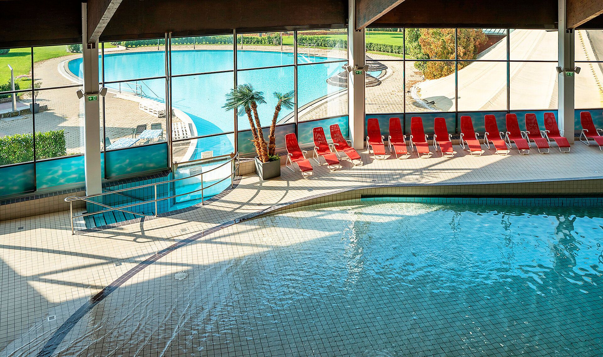 Einladende Sonnenstrahlen durchfluten den Raum und reflektieren sich im glitzernden Wasser des Pools. Die roten Liegen laden zum Entspannen ein, während die üppige Grünfläche im Hintergrund eine friedliche Atmosphäre schafft. Hier kann man die Seele baumeln lassen und die Schönheit der Umgebung genießen.