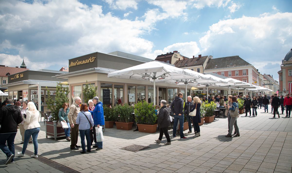 Menschen auf einem belebten Marktplatz mit Cafés und Geschäften unter Sonnenschirmen.