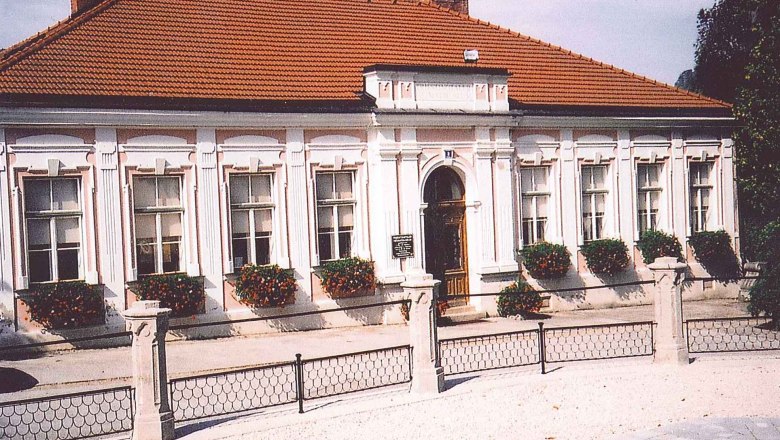 Historisches Geb&auml;ude mit rotem Ziegeldach und wei&szlig;en Fensterrahmen, umgeben von Blumen.