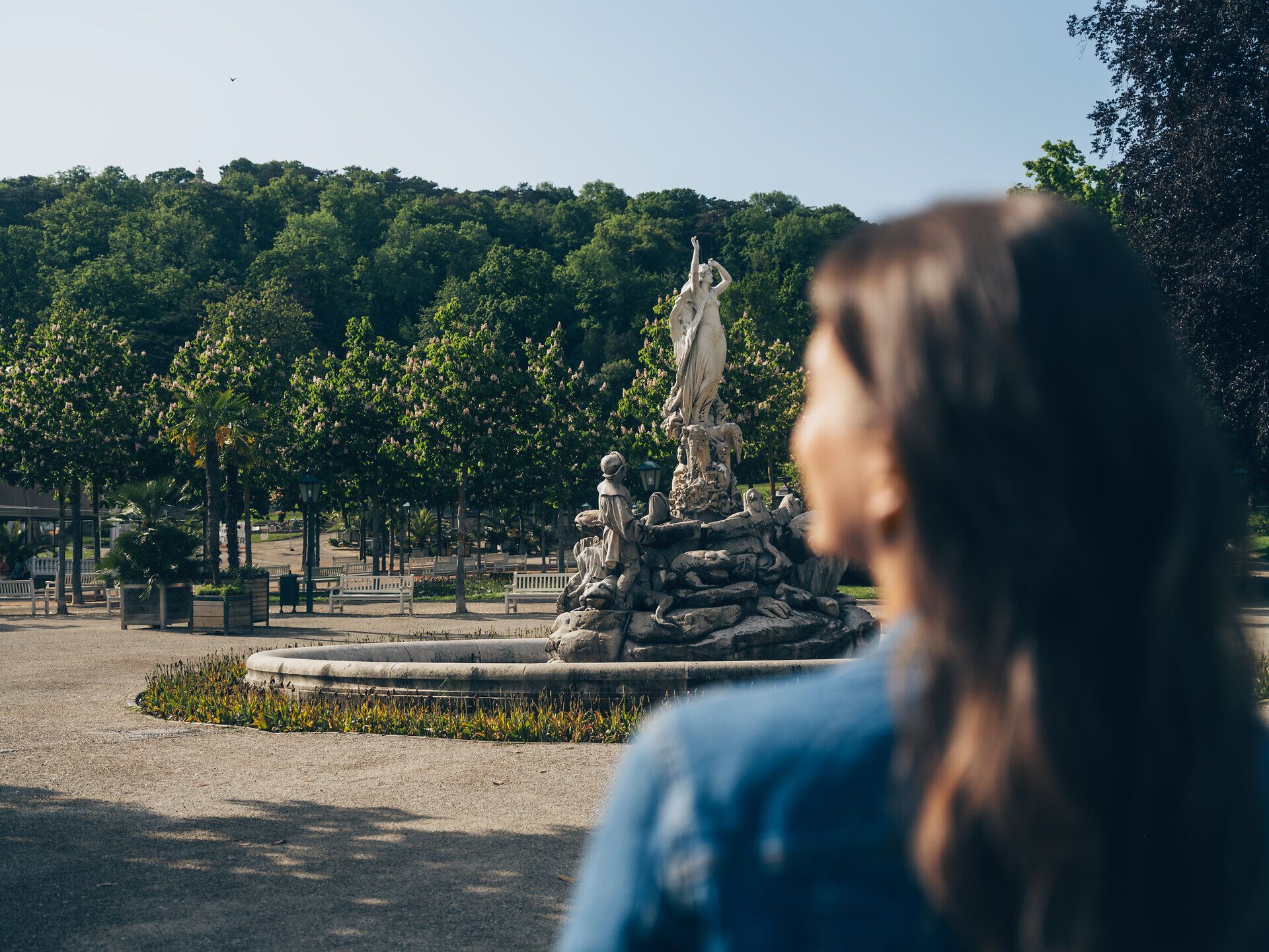Ein sanfter Wind weht durch den Park, während die Sonne die majestätische Statue im Zentrum beleuchtet. Umgeben von üppigem Grün und blühenden Bäumen lädt dieser Ort zum Verweilen und Entspannen ein.
