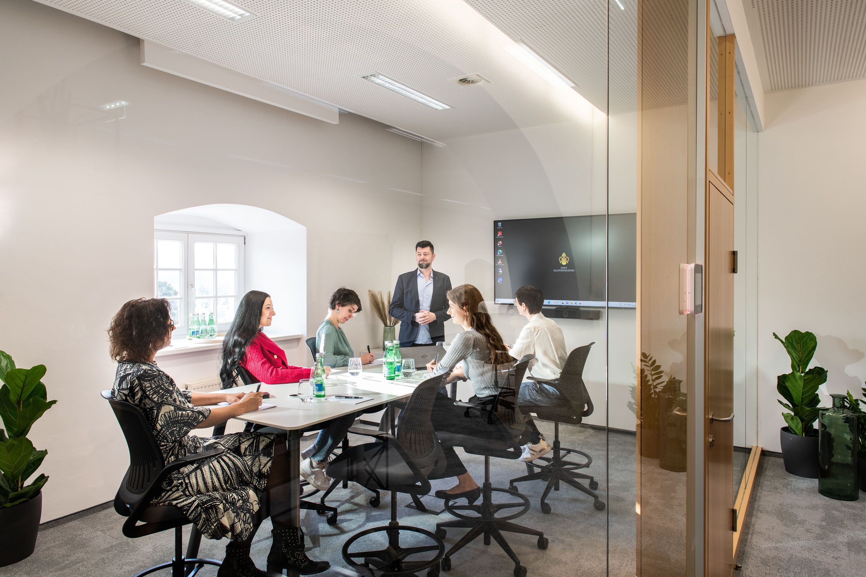 Ein moderner Konferenzraum mit sechs Personen, die an einem Tisch sitzen und einem Vortragenden zuhören. Im Hintergrund ein Bildschirm.