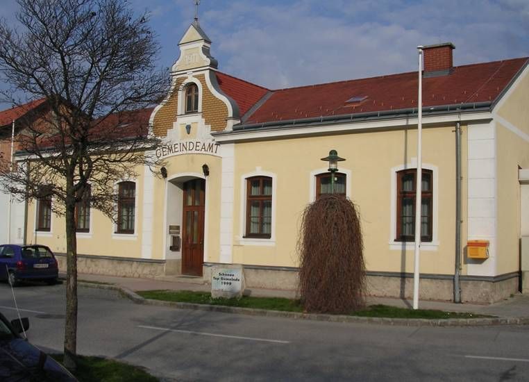Gelbes Gebäude mit der Aufschrift 'Gemeindeamt', rotes Dach, Baum und Laterne davor.