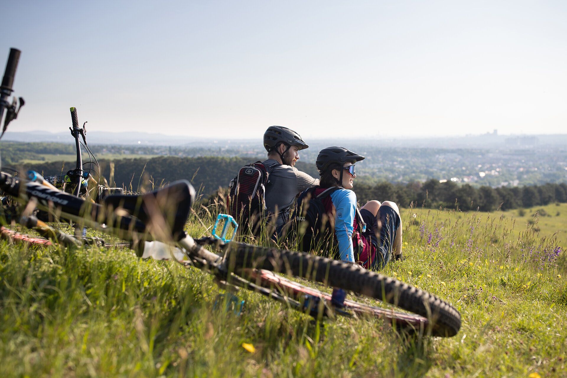 In der warmen Sommerluft genießen zwei Mountainbiker die atemberaubende Aussicht auf die sanften Hügel und das weite Tal. Umgeben von blühenden Wiesen und dem Duft der Natur, ist dies der perfekte Ort für eine wohlverdiente Pause nach einer aufregenden Tour.