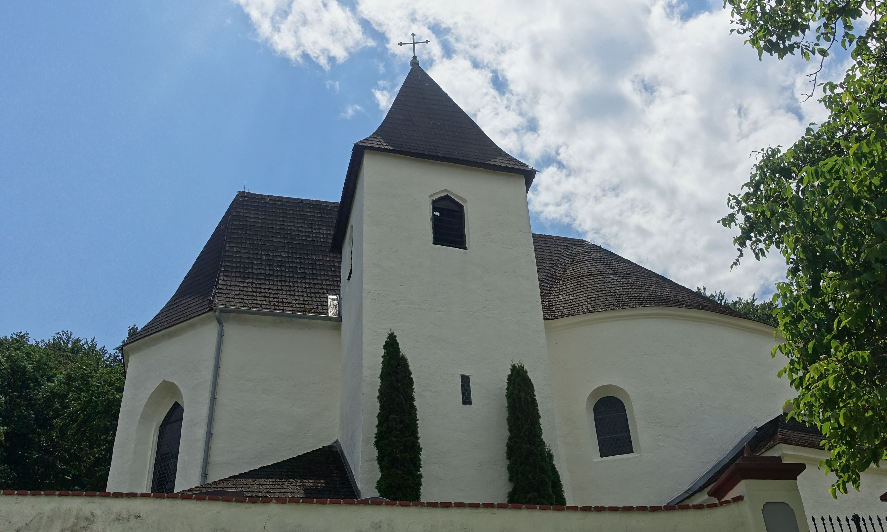Die kleine Rundkirche bei Neulengbach erhebt sich leicht aus der grünen Grasfläche. Mit ihren weiß verputzten Mauern und dem dunklen, steilen Dach wirkt sie schlicht und doch erhaben. Ein schlanker Turm wacht links, während schmalbögige Fenster das Licht lenken. Eingebettet zwischen Bäumen zeigt sie sich als stiller Beobachter der Zeit, ein architektonisches Relikt, das vom Wechselspiel zwischen Natur und Mensch erzählt. Diese versteckte Perle lädt zu einem Ausflug ein, bei dem man die Ruhe und Geschichte inmitten der Natur erleben kann.
