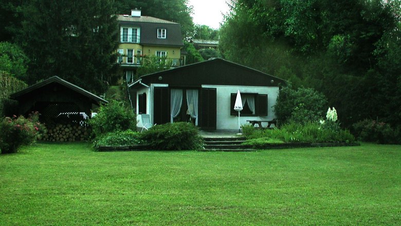 Ein kleines Haus mit Garten und Terrasse, umgeben von Bäumen und einem größeren Gebäude im Hintergrund.