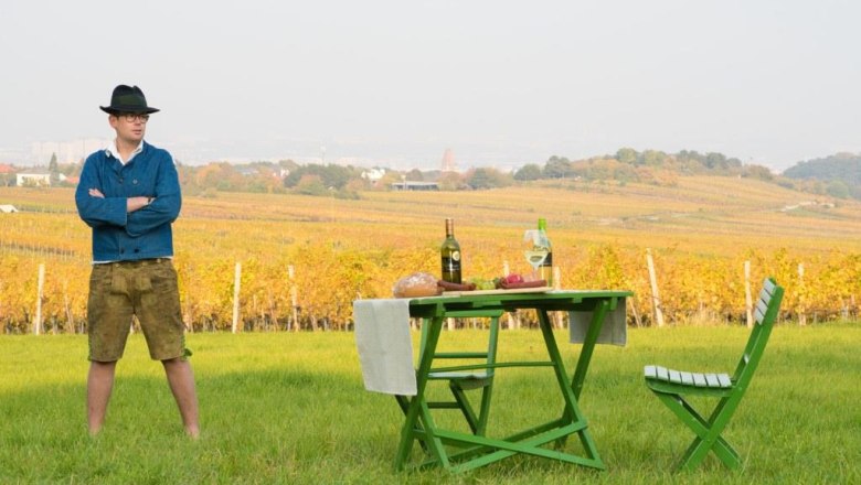 Man in traditional dress stands on a meadow in front of a table with bottles of wine and bread, vineyards in the background.