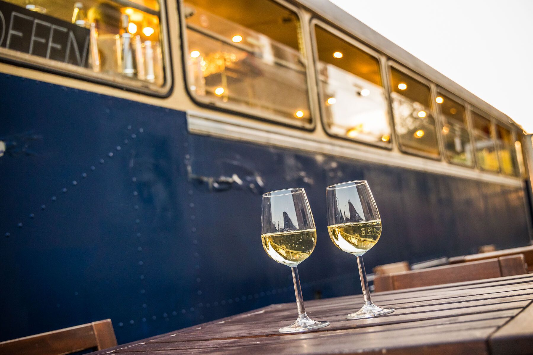 Genießen Sie einen entspannten Abend auf der Terrasse des Kulturbahnhofs, wo zwei Gläser erfrischender Weißwein auf einem rustikalen Holztisch stehen. Die sanften Farben des Sonnenuntergangs spiegeln sich in den Gläsern wider und schaffen eine einladende Atmosphäre, die zum Verweilen einlädt.