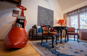 A stylishly furnished hotel room with a large red shoe as a bookshelf, antique chairs, a table and a floor lamp.