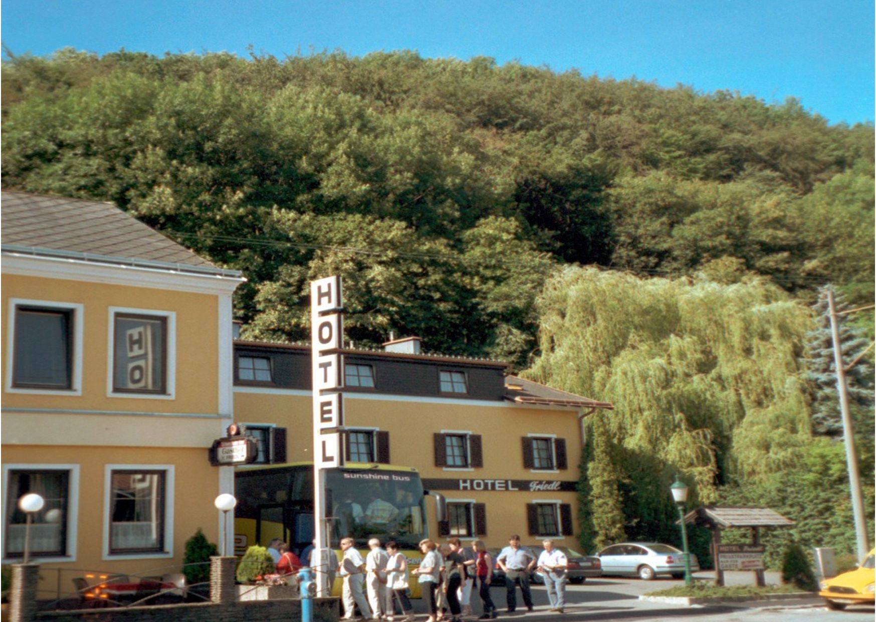 Ein gelbes Hotelgebäude mit einem großen Hotelschild und Menschen davor, umgeben von Bäumen.