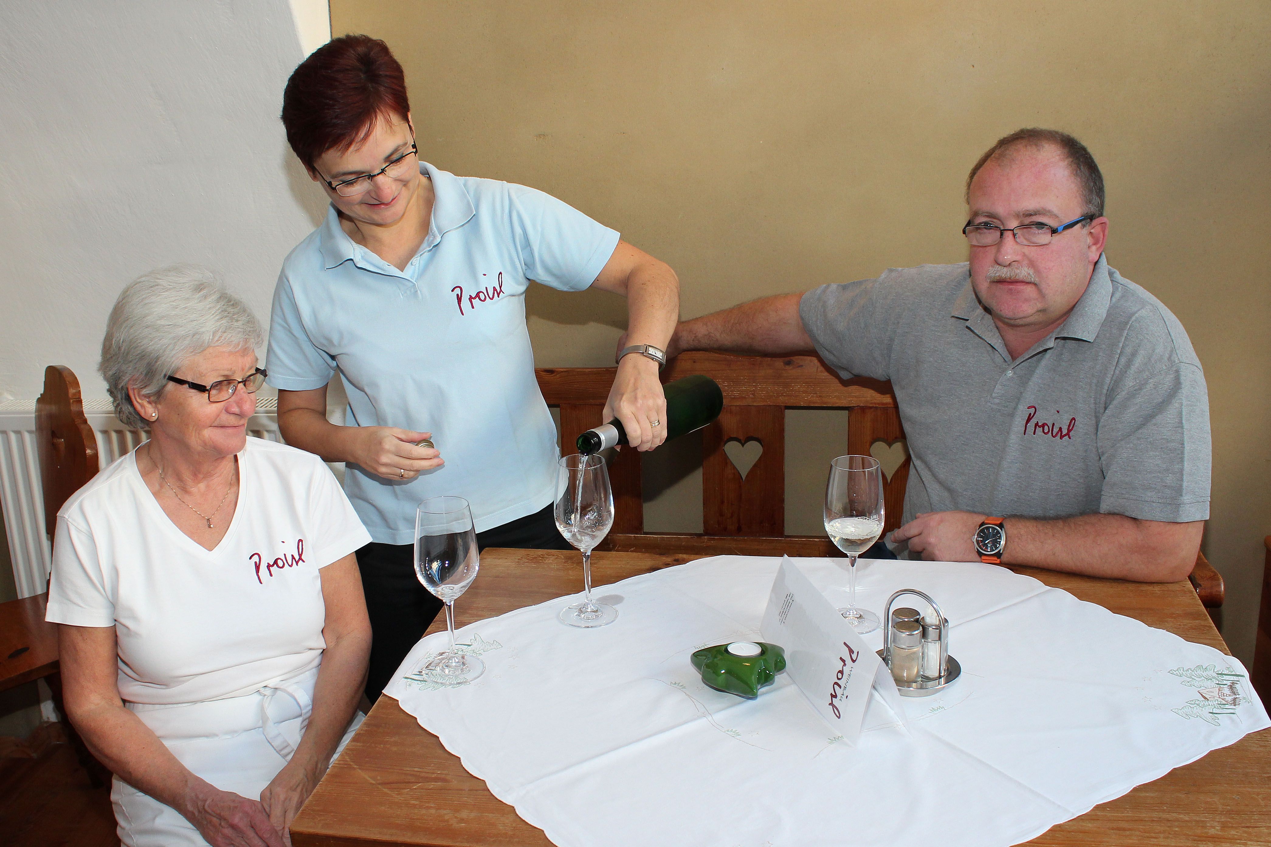 Drei Personen sitzen an einem Tisch, eine Frau schenkt Wein ein.