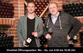 Zwei Männer in einer Weinkellerei, umgeben von Weinflaschen, einer hält ein Glas Wein.