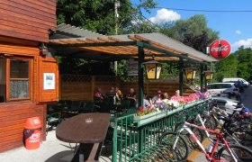 Outdoor area of a snack bar with wooden walls, tables and bicycles. People sitting under a covered area. Coca-Cola sign visible.