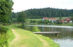 A quiet lake with a jetty, surrounded by green countryside and houses in the background.