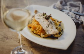 Ein Teller mit Fischfilet auf Nudeln und Gemüse, daneben ein Glas Weißwein.