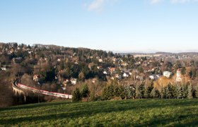 Landschaft mit Zug auf Viadukt in Eichgraben, umgeben von Hügeln und Häusern.