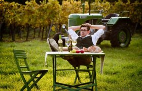 Ein Mann entspannt sich an einem Tisch im Weinberg mit Wein, Brot und &Auml;pfeln. Im Hintergrund steht ein gr&uuml;ner Traktor.