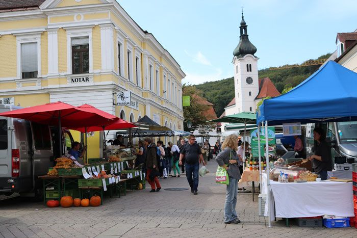 Ein belebter Bauernmarkt mit Ständen und Besuchern vor einem gelben Gebäude und einer Kirche im Hintergrund.