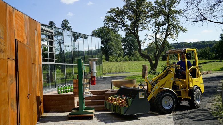 Ein gelber Elektro-Hoftrac transportiert Kisten mit Äpfeln vor einem modernen Gebäude auf einem Bauernhof.