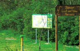 Holzschild mit der Aufschrift 'Blumauer Lauf- und Fitness Parcours' vor einer Infotafel im Grünen.