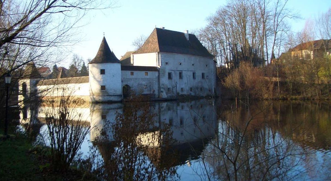 Ein historisches Gebäude mit Türmen spiegelt sich in einem ruhigen Gewässer, umgeben von kahlen Bäumen.