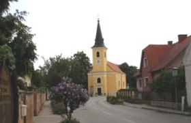 Eine gelbe Kirche mit Turm in einer ruhigen Straße, umgeben von Bäumen und Häusern.