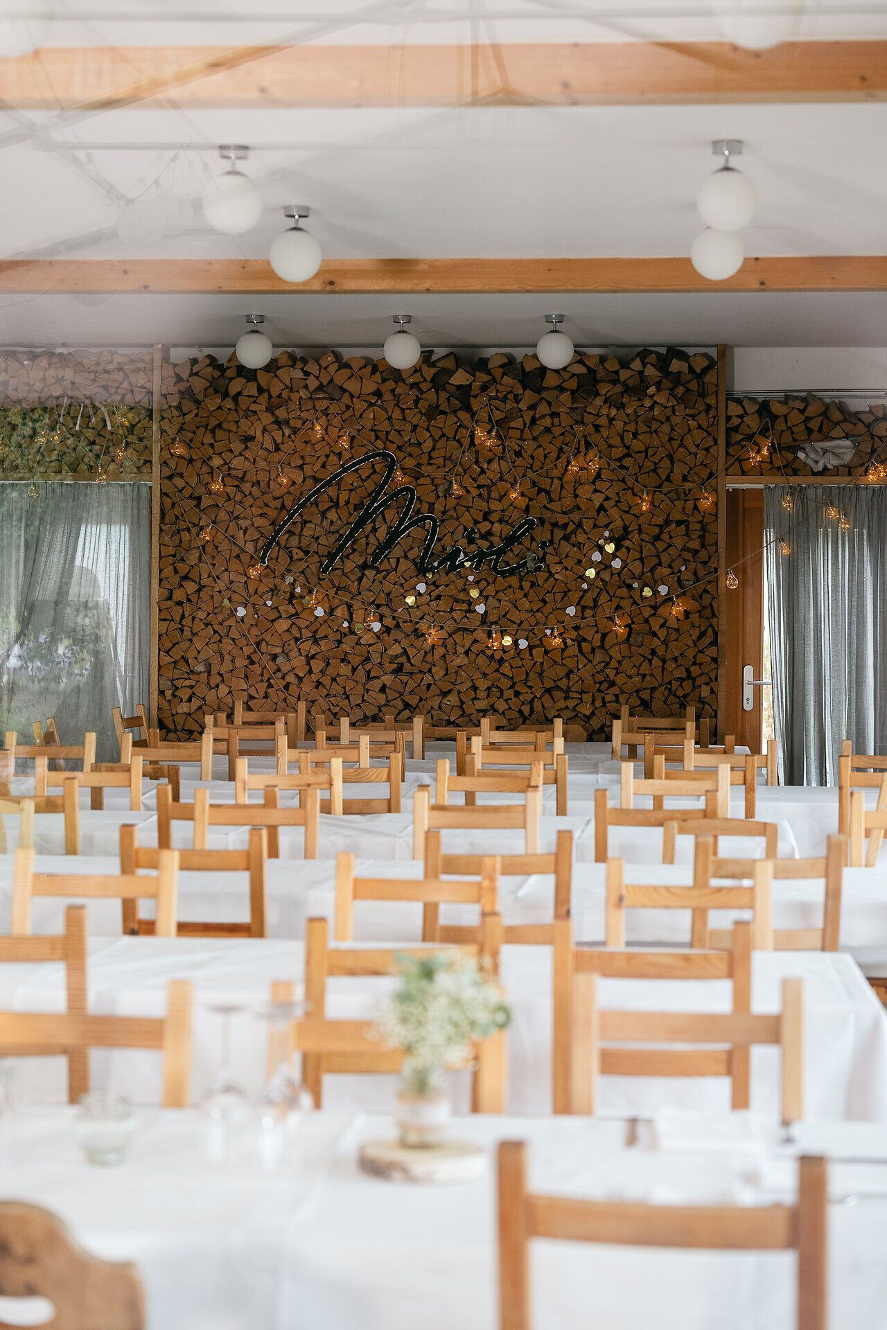 Ein einladender Gastraum mit hellen Holztischen und Stühlen. Im Hintergrund ist eine einzigartige, dekorative Wand aus gestapelten Holzscheiten zu sehen, die den Schriftzug 'Mirli' trägt, und dem Raum eine warme, gemütliche Atmosphäre verleiht.