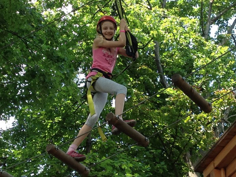 Ein Kind mit Helm und Kletterausrüstung balanciert auf einem Hochseilparcours zwischen Bäumen.