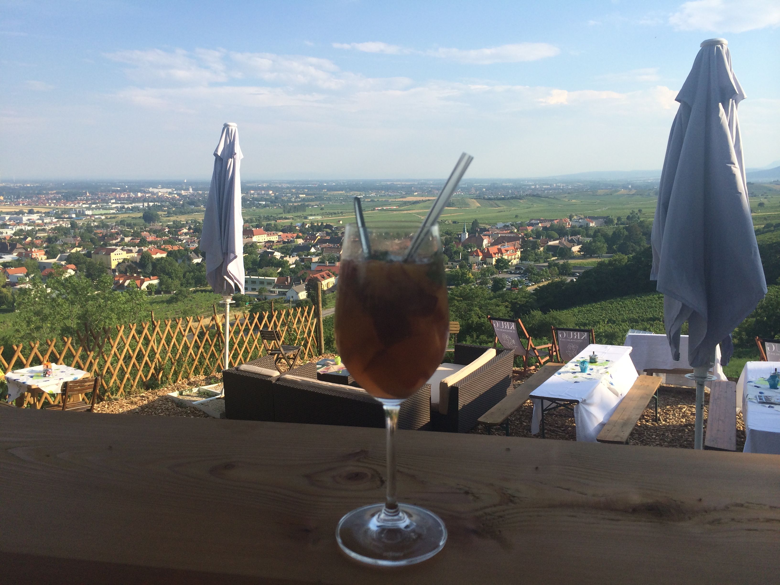 Ein Getränk in einem Weinglas mit Strohhalmen vor einer Landschaft mit Tischen und Sonnenschirmen.