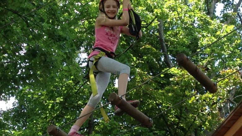 A child wearing a helmet and climbing equipment balances between trees on a high ropes course.