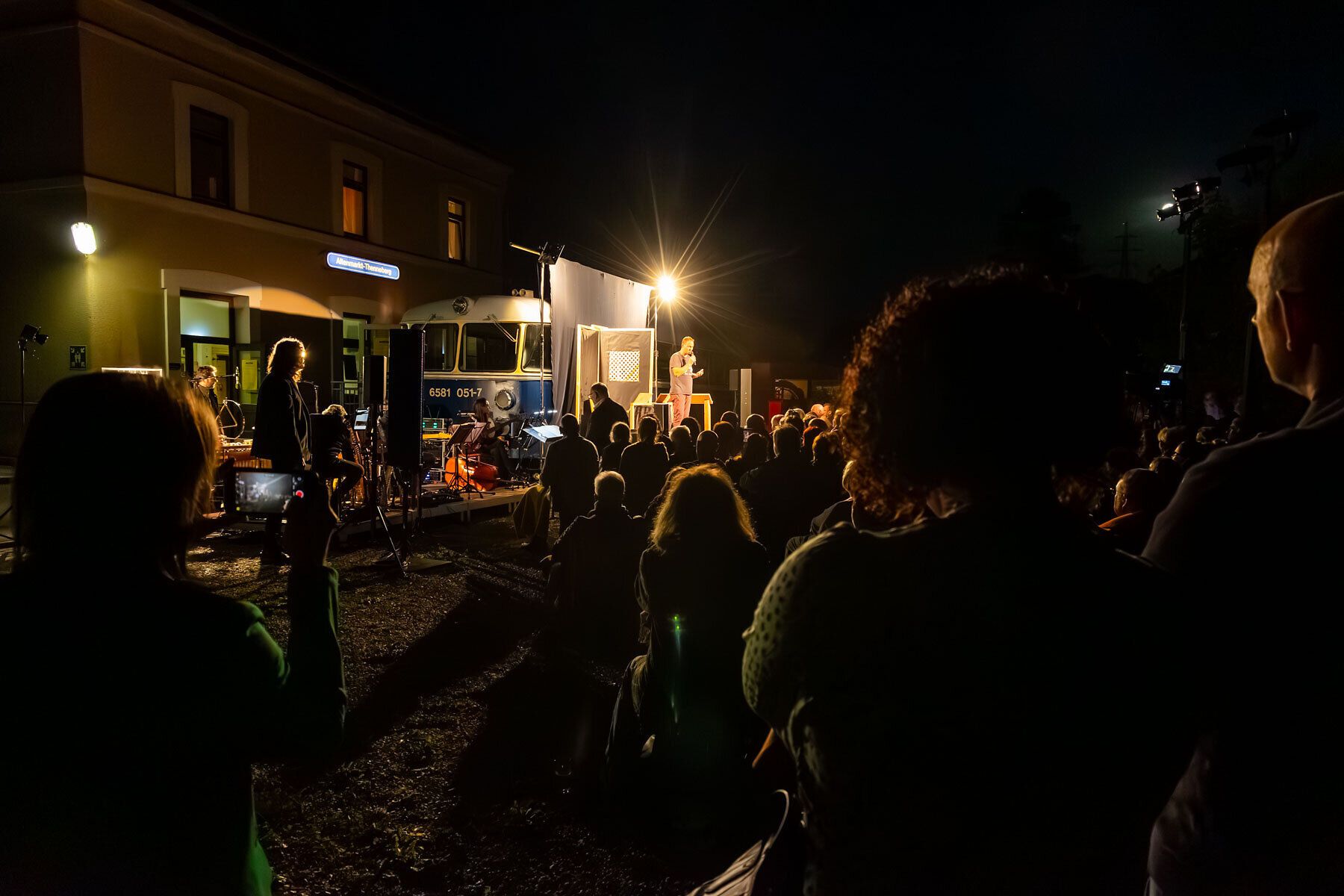 Unter dem strahlenden Licht der Bühne versammeln sich Menschen, um die magische Atmosphäre eines kulturellen Abends zu genießen. Die Klänge der Darbietung vermischen sich mit dem sanften Rascheln der Blätter, während die Nacht langsam hereinbricht und die Umgebung in ein warmes Licht taucht.