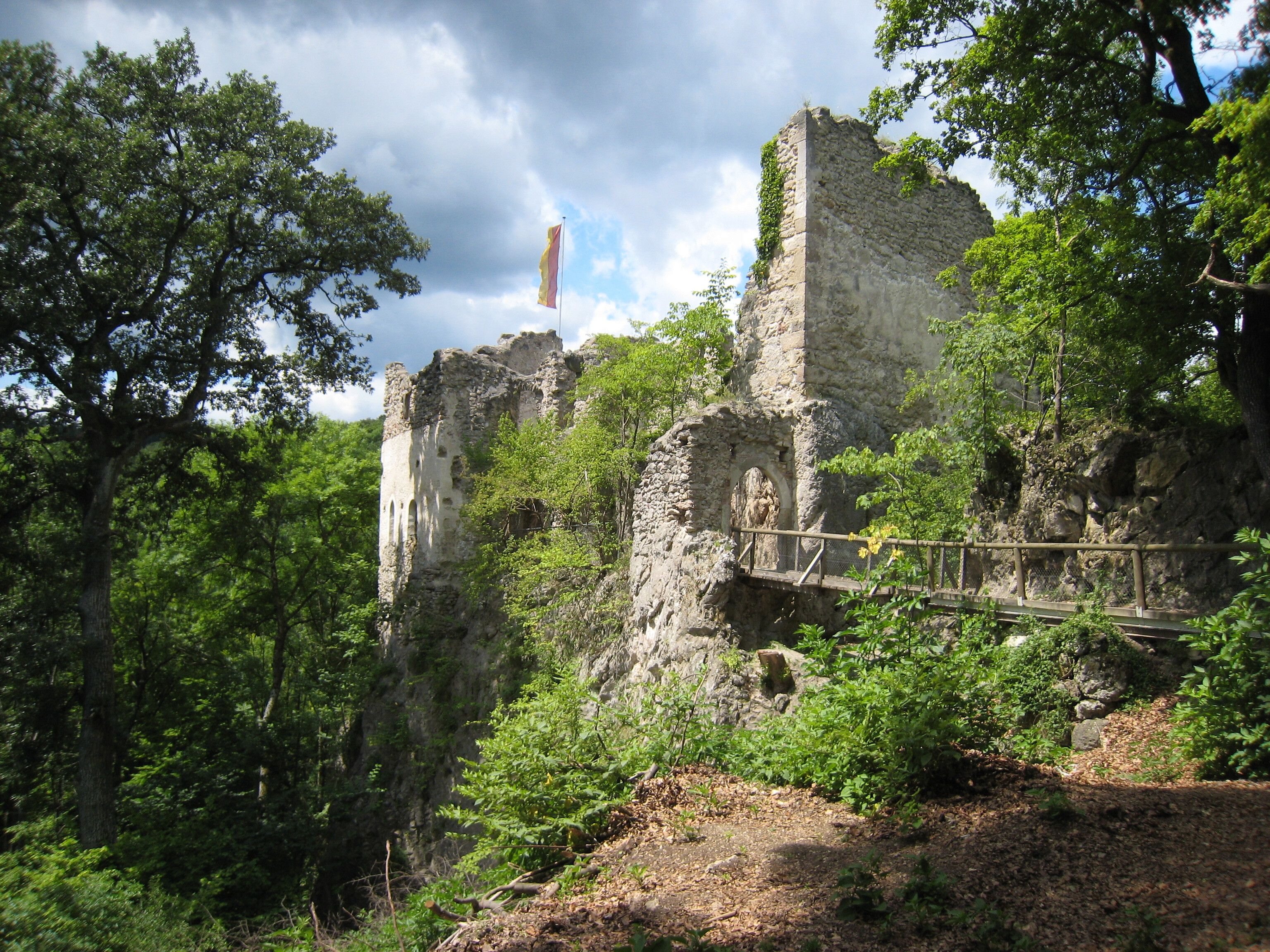 Die Ruine, umgeben von üppigem Grün und alten Bäumen, strahlt eine geheimnisvolle Atmosphäre aus. Der Blick auf die verwitterten Mauern und die sanften Hügel lädt dazu ein, in die Geschichte einzutauchen und die Schönheit der Natur zu genießen.