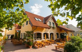 Ein gelbes Gebäude mit roten Dachziegeln und einem Biergarten mit Holzbänken und Sonnenschirmen.