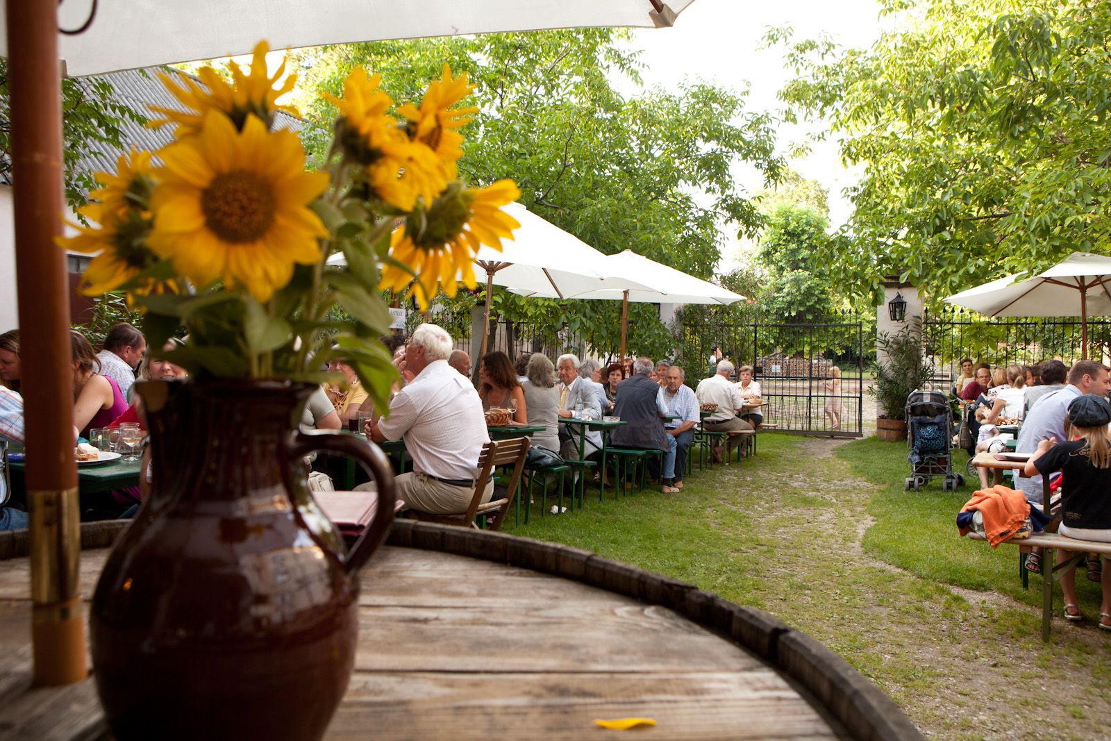 Ein Biergarten mit Menschen an Tischen, Sonnenschirmen und Sonnenblumen in einem Krug im Vordergrund.