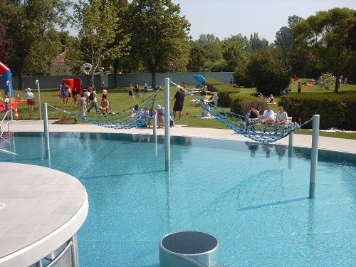 An outdoor pool with hammocks over the water and people in the background on the sunbathing lawn.