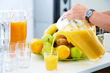 Person gießt Orangensaft aus einem Krug in ein Glas, umgeben von frischem Obst.