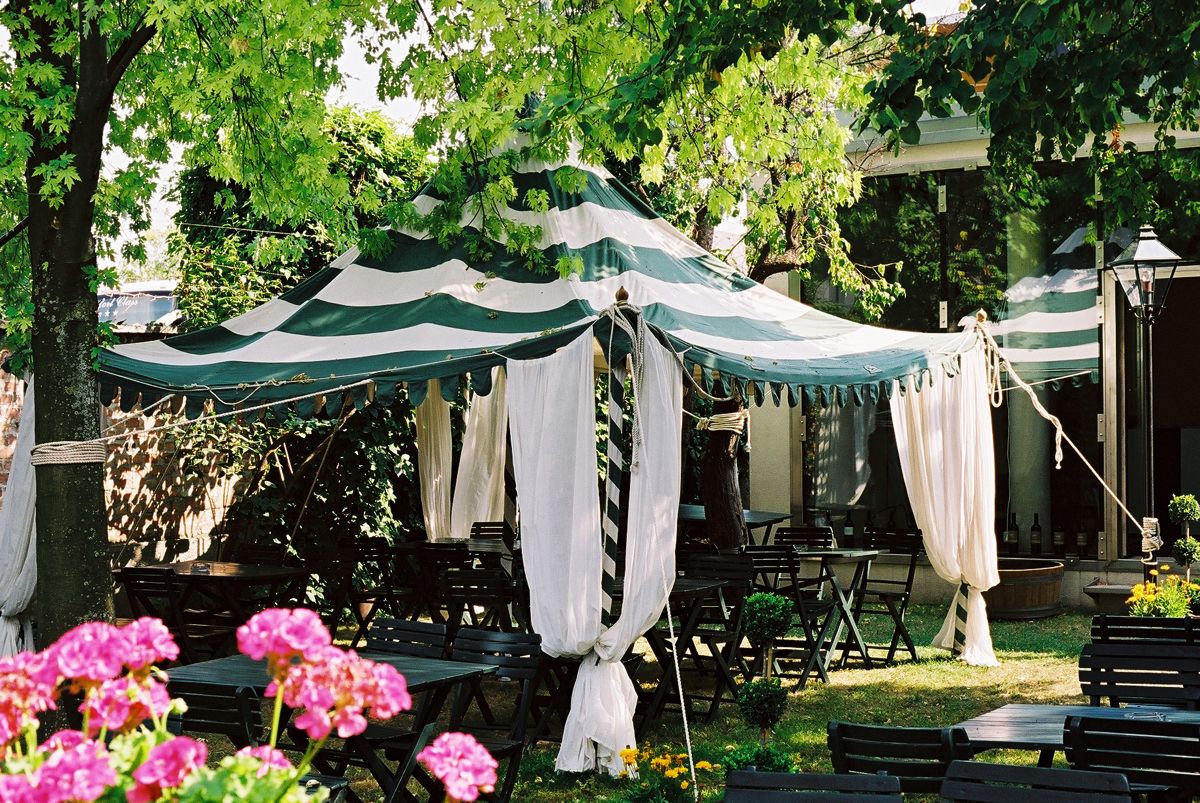 Ein gestreiftes Zelt mit weißen Vorhängen steht in einem Garten, umgeben von Bäumen und Blumen.