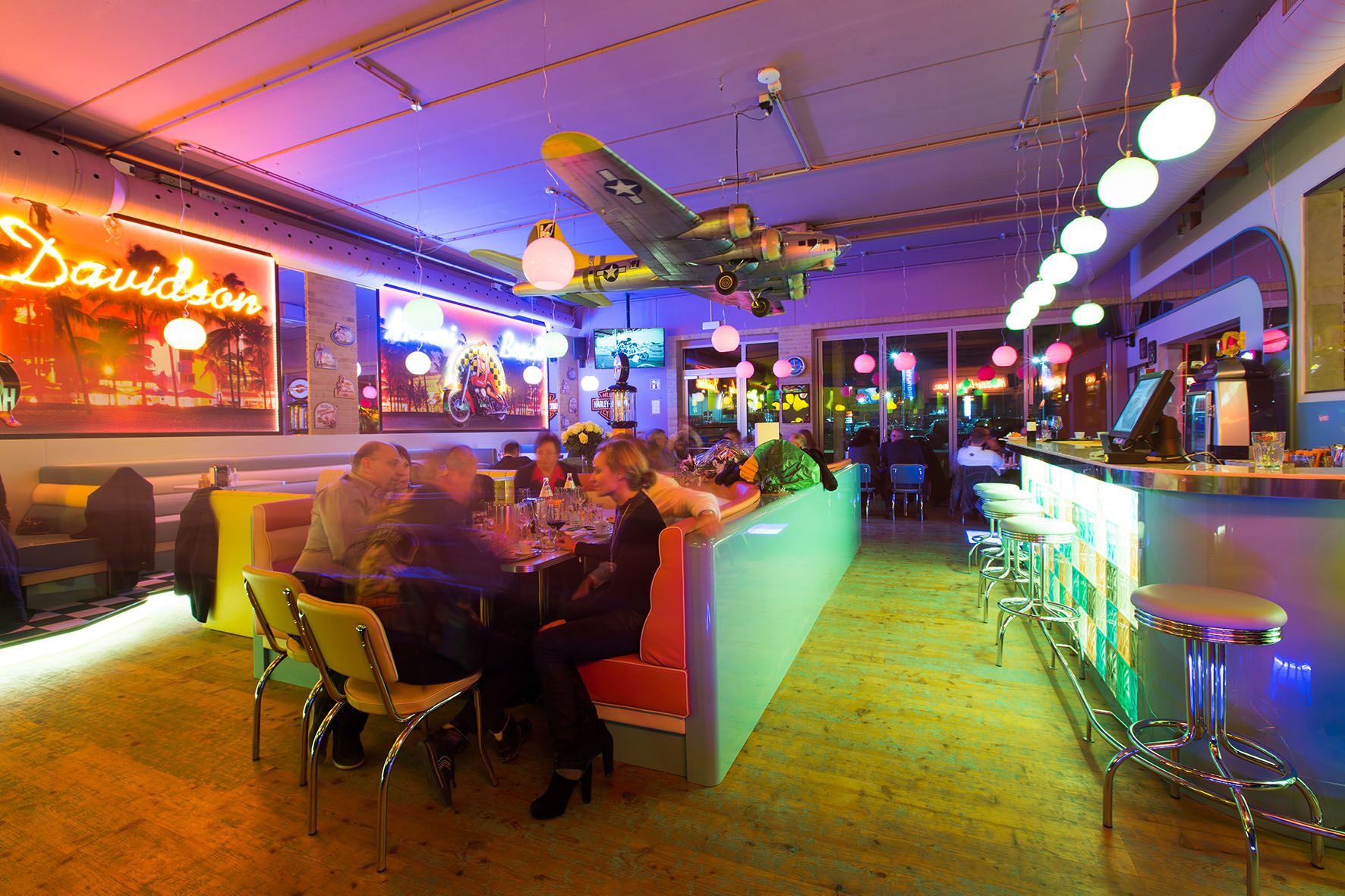 Innenansicht eines bunt beleuchteten Diners mit Bar, Tischen und einem Flugzeugmodell an der Decke.