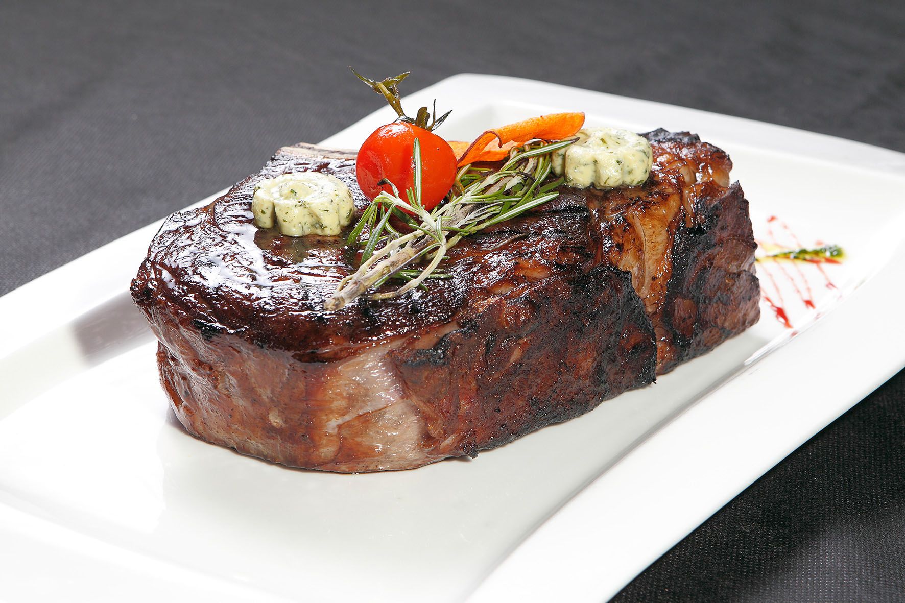 Ein saftiges Steak mit Kräuterbutter, Rosmarin und einer Kirschtomate auf einem weißen Teller.
